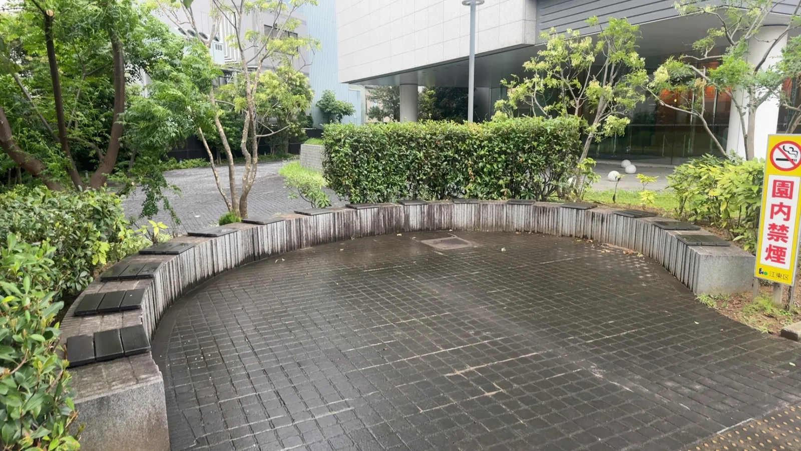 曲線ベンチと生け垣、園内禁煙の黄色い看板。ビル前の小さな広場