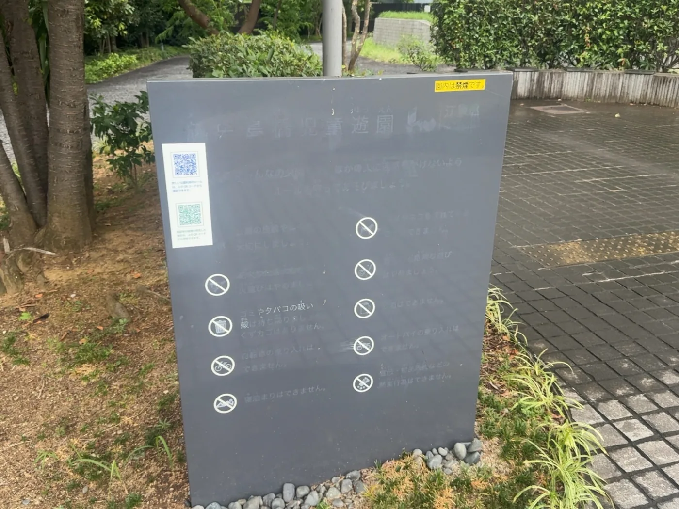 越中島橋児童遊園と書かれた園内の利用案内板。点字ブロックと植栽が見える
