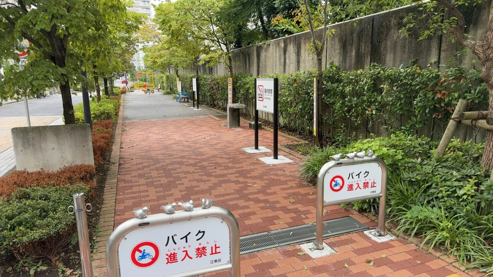 入口の金属ゲートに雀の小さな置物。レンガ敷き、園内禁煙の看板、奥に水飲み場と遊具