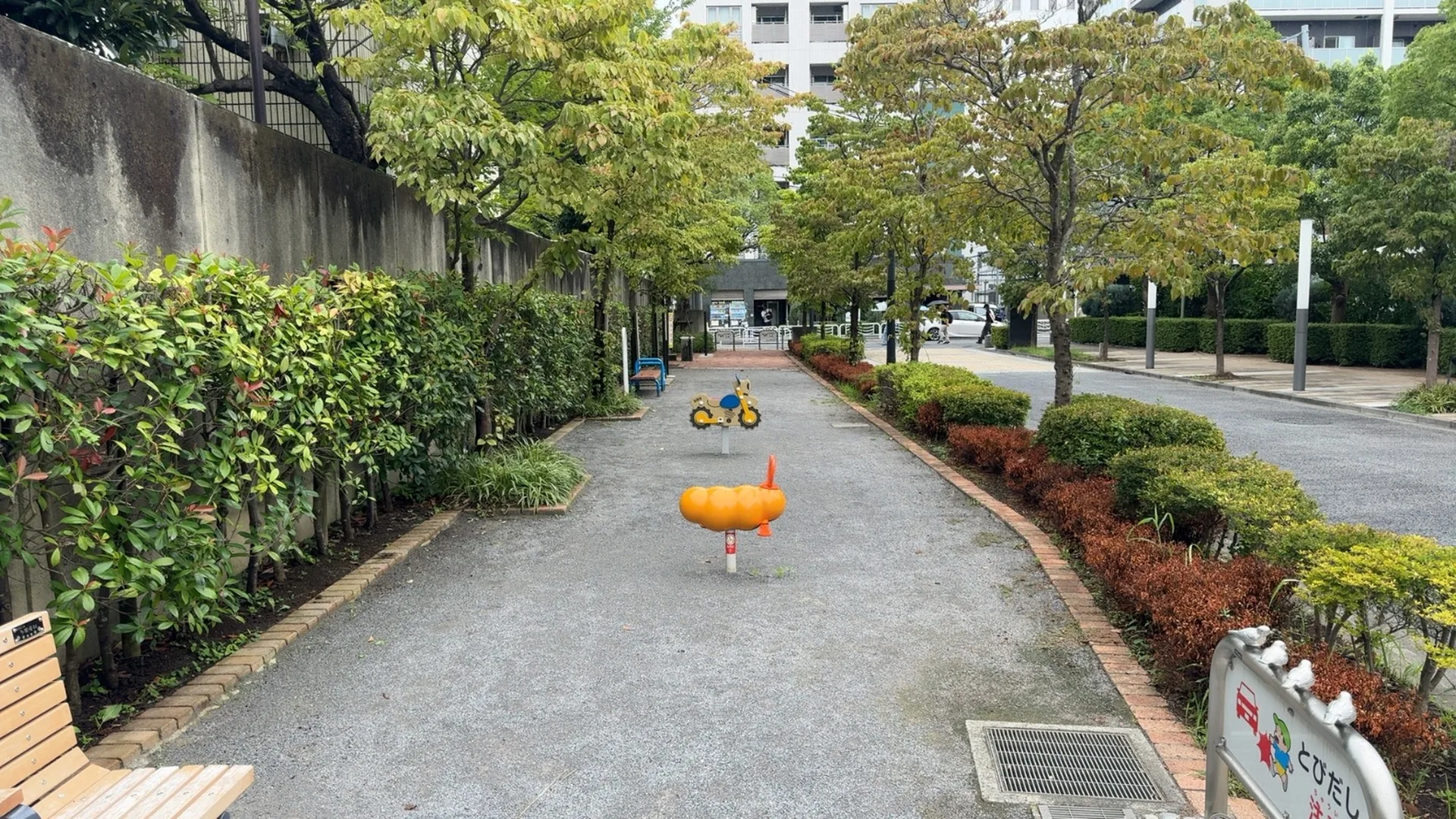細長い砂利の園路に、黄色いバイク型とオレンジ色のイモムシ型スプリング遊具が並ぶ新砂一丁目児童遊園