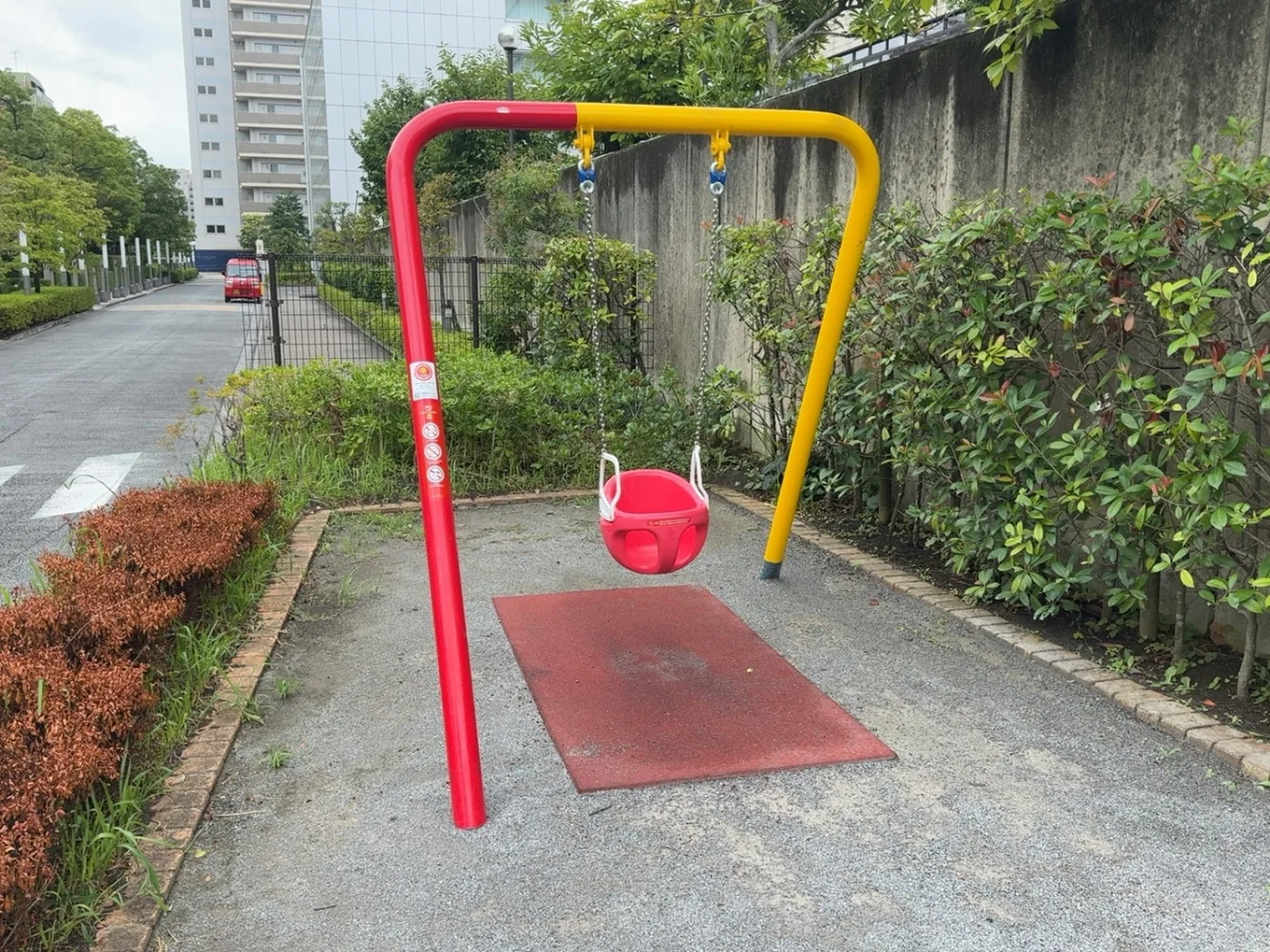 赤と黄のフレームに赤いバケツ座席が下がる幼児用ブランコ。足元に赤い安全マット