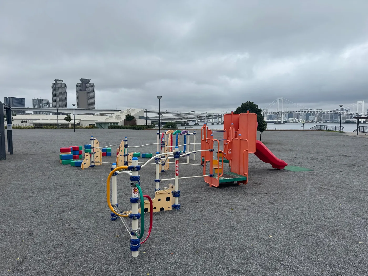 赤い滑り台とオレンジのパネルがある遊具。奥にレインボーブリッジ